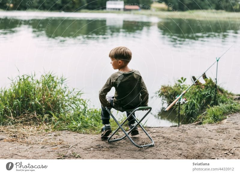 Der Junge sitzt auf einem Klappstuhl am Ufer eines Sees oder Flusses. Erholung, Wochenenden, Tourismus. Rückansicht Stuhl Kind Sommer Lifestyle Freizeit