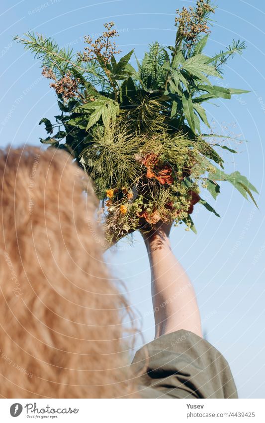 Schöne Frau mit einem Blumenstrauß aus Wildblumen. Eine hübsche blonde Frau mittleren Alters in einem grünen Kleid mit lockigem Haar. Blauer Sommerhimmel im Hintergrund. Rückansicht. Vertikale Aufnahme