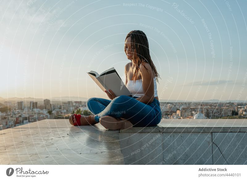 Coole schwarze Frau, die auf einem Dach in der Stadt ein Buch liest lesen Lehrbuch freie Zeit Literatur trendy Beine gekreuzt achtsam Dachterrasse Großstadt