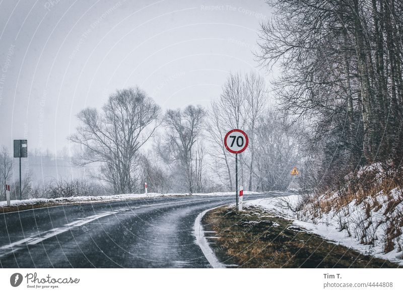 die Kurve einer Landstraße im Winter. Im Vordergrund ein Verkehrsschild 70 Polen Straße Schnee Außenaufnahme kalt Menschenleer Baum Landschaft Natur