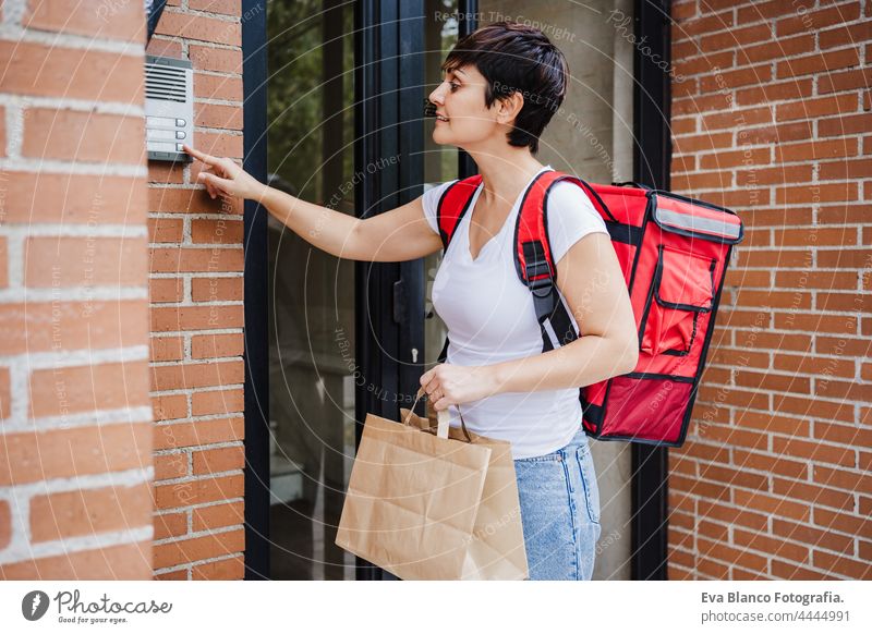 Junge Reiterin mit rotem Rucksack, die eine Pappschachtel mit Lebensmitteln in der Stadt hält. Konzept eines Lieferdienstes. Frau benutzt eine Gegensprechanlage