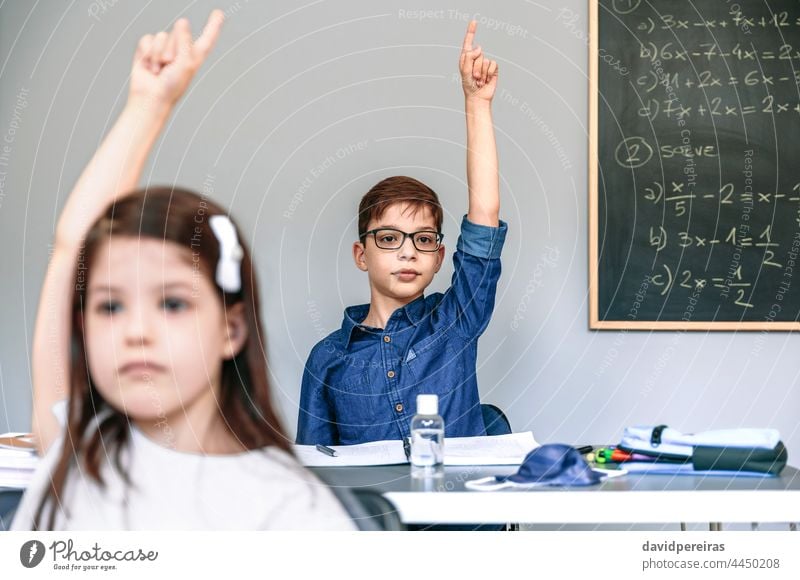 Schüler mit Maske auf dem Tisch heben die Hände in der Schule neue Normale Coronavirus Handreichung Klasse teilnehmend Sicherheit Virus Menschen Mädchen Junge