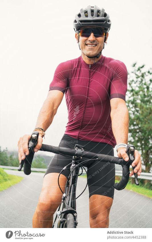 Lächelnder Radfahrer mit Schutzhelm auf dem Fahrrad auf der Fahrbahn Sport Fahrradfahren Sonnenbrille Mann Landschaft Porträt kreativ Design Straße behüten