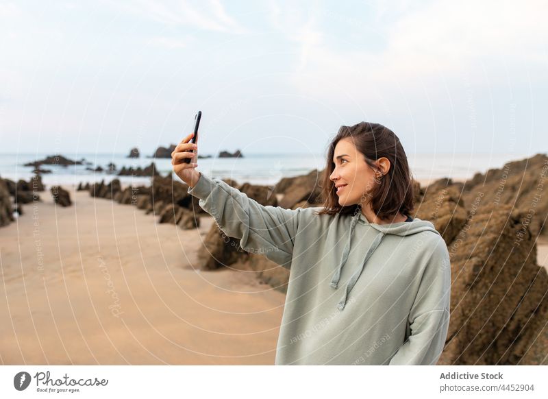 Lächelnde Frau, die ein Selfie mit ihrem Smartphone am felsigen Strand macht Selbstportrait Meeresküste Felsen Gedächtnis Moment Himmel Freizeit benutzend