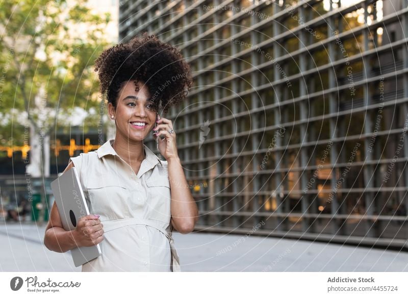 Lächelnde schwarze schwangere Frau, die mit einem Smartphone spricht und einen Laptop trägt benutzend reden Gespräch positiv führen Bauch warten Talkrunde