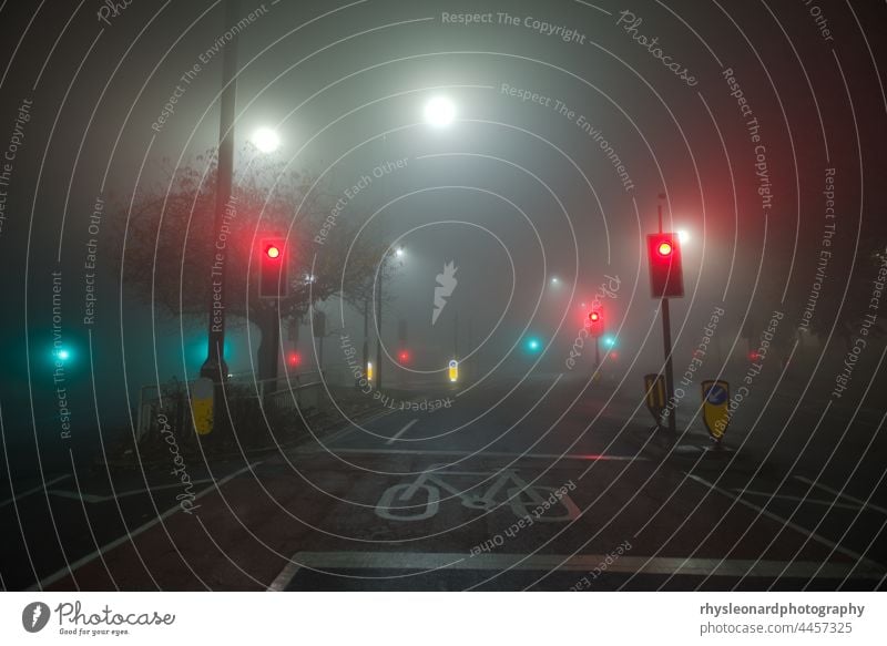 Bunte Ampeln und weiße Straßenlaternen leuchten durch dichten Nebel. An einer Kreuzung von zwei Bundesstraßen in Sheffield. Es ist Nacht, die Straßen sind also völlig leer oder menschenleer.