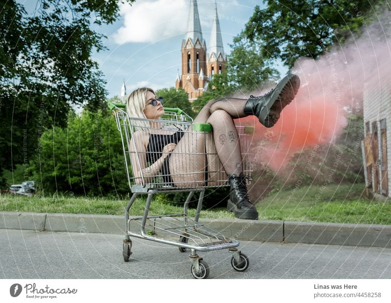 Was für ein wunderschönes blondes Mädchen ist chillen auf dem Parkplatz. Roter Rauch, hübsche lange Beine, Sonnenbrille, schwarze Stiefel, und eine modische schöne Frau auf dem Bild. Entspannen Sie sich und genießen Sie das perfekte Wetter wie niemand sonst.