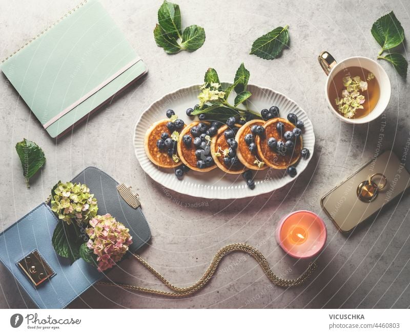 Blaubeeren Pfannkuchen Frühstück auf Home-Office-Schreibtisch mit Tasse Tee, Handtasche, Blumen, Smartphone, Duftkerze, und Hortensienblüten. Ansicht von oben