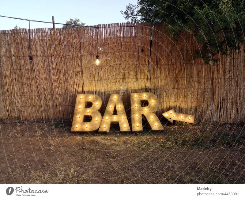 Beleuchteter Hinweis auf eine Bar in Naturfarben vor farblich passendem Reisigzaun im Sommer in der Dämmerung bei einer türkischen Hochzeit im Park in Güzelbahce in der Provinz Izmir am Ägäischen Meer in der Türkei