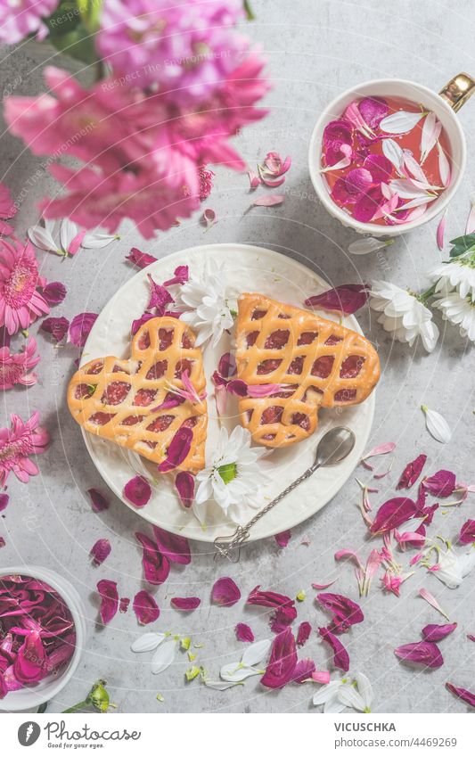 Zwei Herzkuchen auf hellem Tisch mit Tasse rosa Blumentee, Pfingstrosenstrauß und vielen Blütenblättern . Ansicht von oben zwei Kuchen Licht Haufen Draufsicht