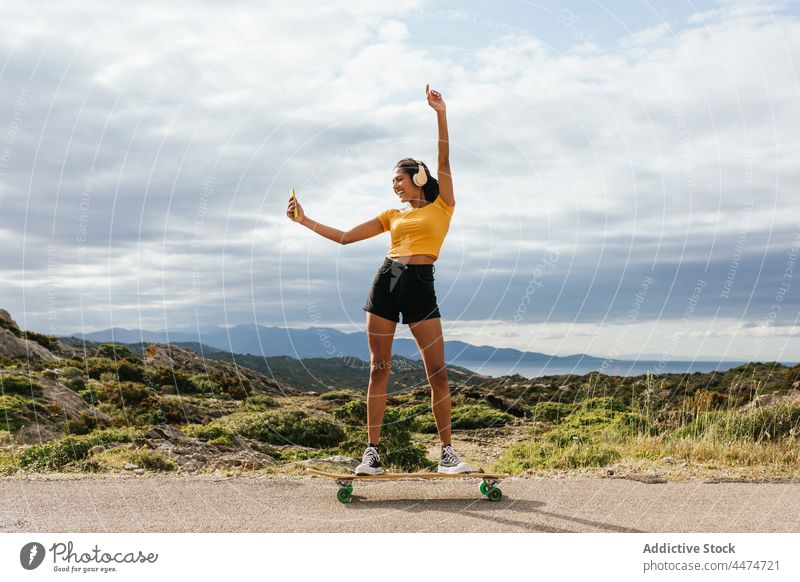 Lächelnde ethnische Frau auf Longboard hebt die Hand und macht ein Selfie Glück Kopfhörer zuhören benutzend Musik Smartphone Freizeit Optimist Aktivität Hipster