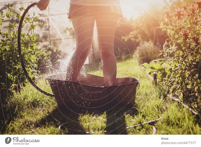 Baden im Sommergarten, Frau, die sich Wasser aus einem Gartenschlauch auf die Füße gießt, stehend in einer Eisenwanne auf einem Gartenweg im Sonnenlicht