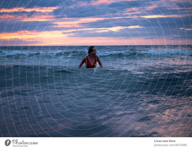 Was für ein herrlicher rot-rosa-violetter Himmel an einem perfekten Abend am Meer. Eine hübsche Frau in einem roten Badeanzug genießt die Wellen. Verlockend blaues Wasser überall, wo man hinsieht. Ein weibliches Model im Meer ist allein, fühlt sich aber nicht einsam.