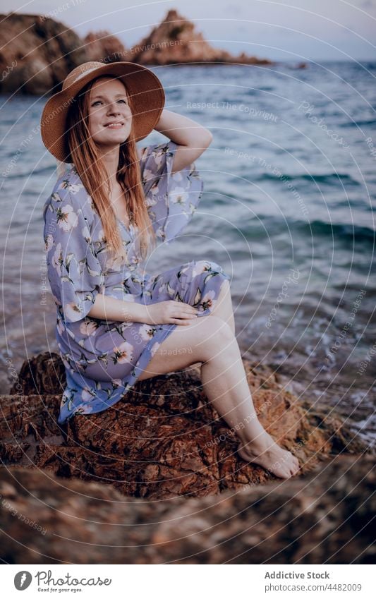 Stilvolle Frau sitzt am Meeresufer MEER Abend Strand Küste Sommer Felsen Kleid Hut Sonnenuntergang charmant Natur Wasser jung Ufer Stein verträumt Windstille