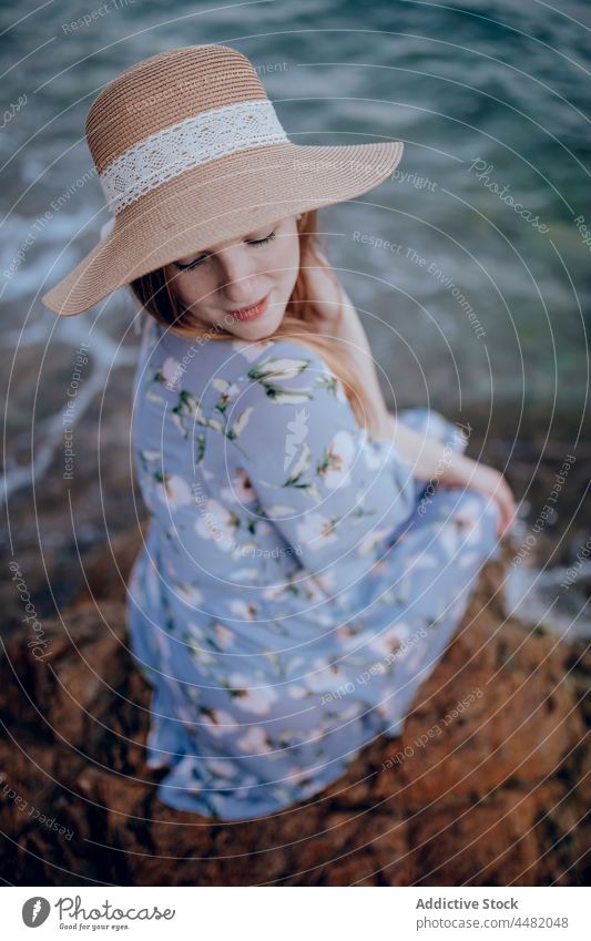 Stilvolle Frau sitzt am Meeresufer MEER Abend Strand Küste Sommer Felsen Kleid Hut Sonnenuntergang charmant Natur Wasser jung Ufer Stein verträumt Windstille