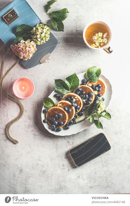 Femininer Frühstücks-Lifestyle mit Blaubeer-Pfannkuchen, Tasse Tee, brennender Kerze und Smartphone mit schwarzem Bildschirm, blauer Handtasche mit Hortensie auf hellgrauem Betontisch. Ansicht von oben.