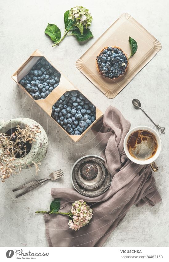 Flat Lay mit Blaubeeren Kuchen, Kaffee, Holzkorb mit frischen Blaubeeren, Geschirrtuch, Wasserglas und Blumen auf weißem Hintergrund. Gemütliche Teezeit mit hausgemachtem Gebäck und heißen Getränken. Ansicht von oben.