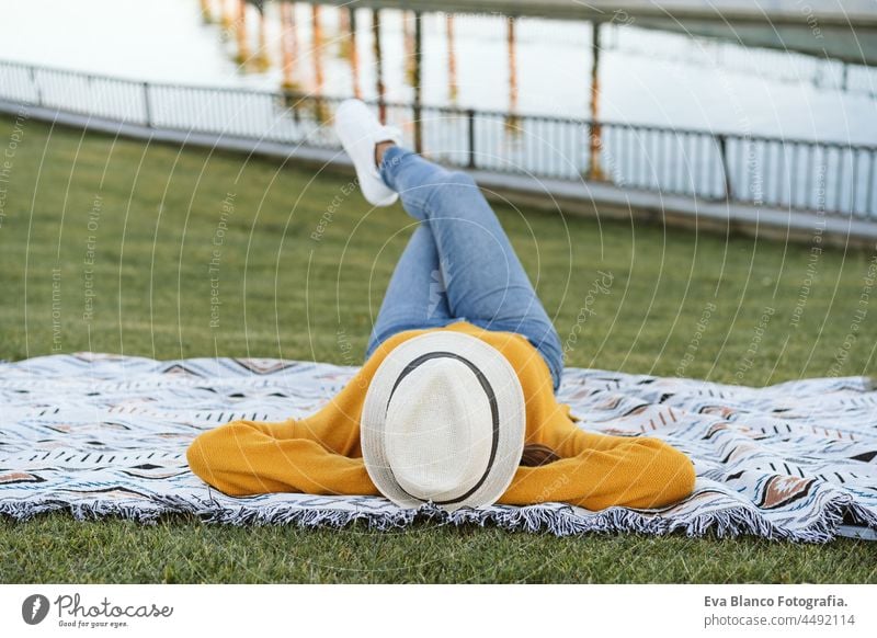 Rückenansicht einer im Gras liegenden Frau mit Hut und gelbem Pullover. Gekreuzte Beine. Entspannung im Freien in einem Park mit See. Lebensstil Lügen