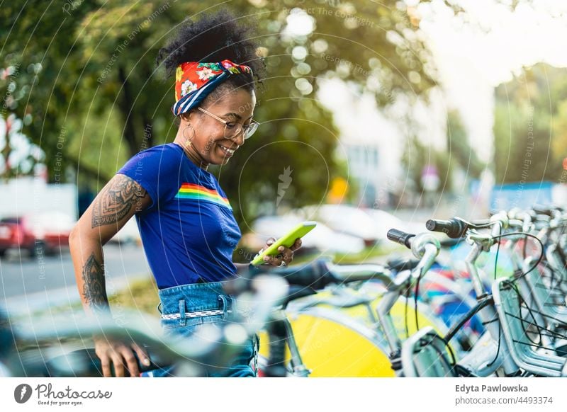 Junge Frau benutzt ein Mietfahrrad in der Stadt mit Smartphone Jahrtausende Brille lockig Freude außerhalb farbenfroh Afro-Look selbstbewusst Schönheit Urlaub