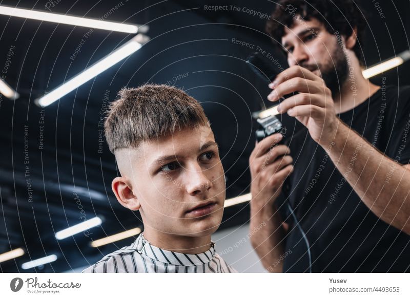 Stilvolle bärtige brünette Friseur macht einen modischen Haarschnitt zu einem jungen Mann. Menschen bei der Arbeit. Kleine Unternehmen oder Dienstleistungen Konzept. Professionelle Haarschnitt und Styling in einem Friseurladen oder Friseursalon