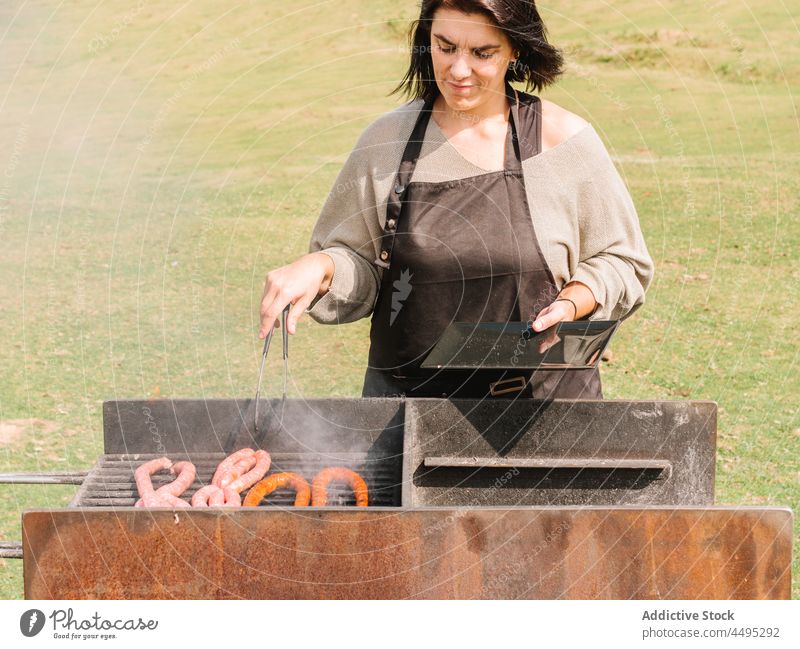 Frau brät Würstchen auf dem Grill auf dem Lande Koch Wurstwaren Fleisch Grillrost Barbecue Braten Landschaft Gitter Feld grillen Lebensmittel geschmackvoll