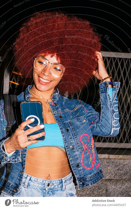 Positive Frau, die auf einer dunklen Straße auf ihrem Smartphone surft Browsen online Afro-Look Abend urban Mode Stil trendy Bekleidung Surfen Textnachricht