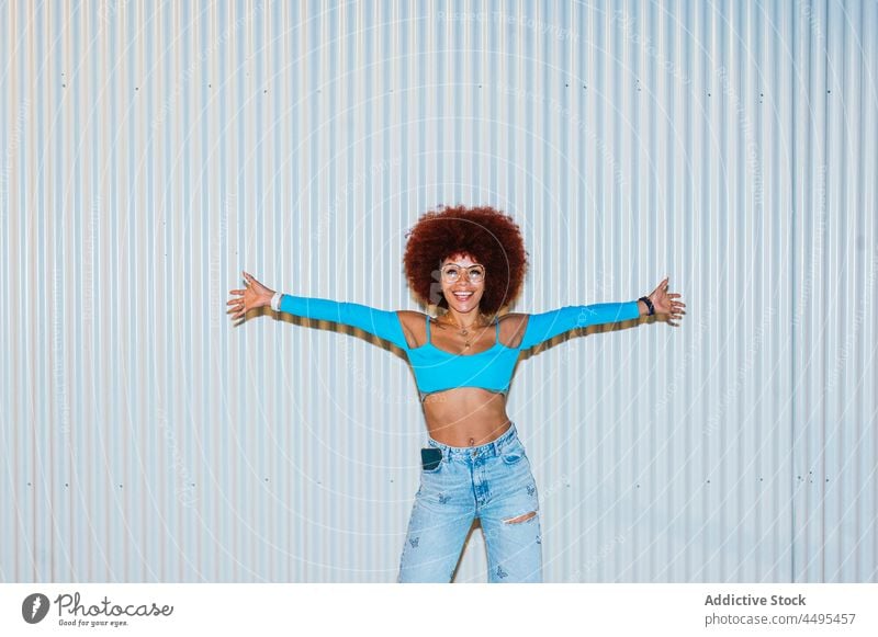 Modische lächelnde Frau mit Afrofrisur steht in der Nähe der Wand Afro-Look Stil Mode trendy Bekleidung Vorschein Outfit Straße Frisur Sonnenbrille Accessoire