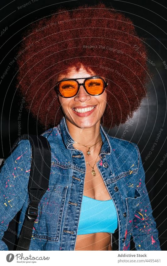 Glückliche Frau in stilvoller Kleidung auf dunkler Straße Jeansstoff Afro-Look Stil Mode Bekleidung Streetstyle Rucksack Abend anhaben dunkel Dämmerung Frisur