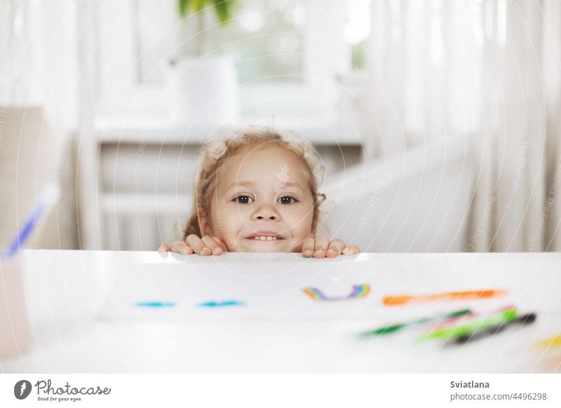 Ein kleines Mädchen versteckt sich unter dem Tisch und schaut darunter hervor. Das Mädchen spielt in der Pause zwischen den Hausaufgaben. Hausaufgaben, Homeschooling, Online-Lernen