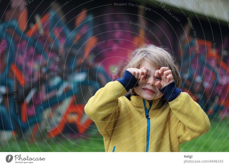 Kind in gelber Jacke macht Faxen vor einer Wand mit Graffitis Junge Oberkörper Wiese Mensch Kindheit Außenaufnahme Farbfoto Porträt 3-8 Jahre