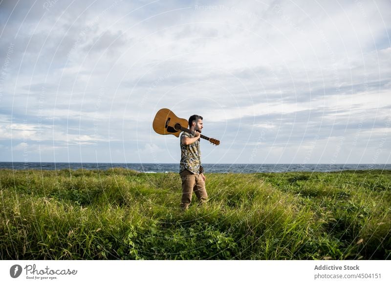 Musiker mit Gitarre am grasbewachsenen Ufer vor wogendem Meer stehend Mann spielen akustisch Gras Instrument Gitarrenspieler männlich Tageslicht Küste grün