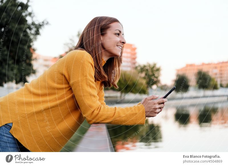 Lächelnde kaukasische Frau mit gelbem Pullover, die ein Mobiltelefon im Freien im Park bei Sonnenuntergang benutzt. Technologie und Lebensstil Handy Sprechen