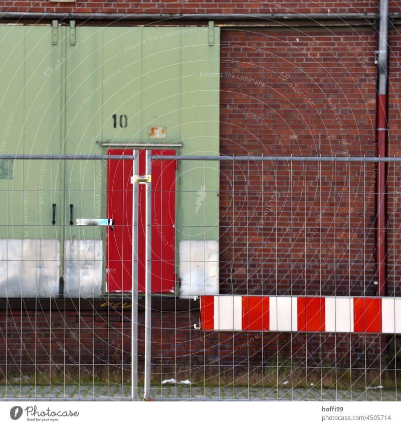 Warnschild an Bauzaun vor Rolltor mit roter Tür an Lagerhalle mit Fallrohr und roten Ziegeln 10 Absperrung weiß Barke Barriere Metall Zaun Gitter rot-weiß Linie