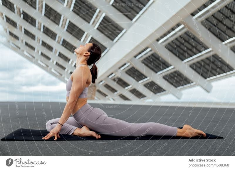 Sportlerin macht halbe Taube Asana in der Nähe von Sonnenkollektoren Frau Halbtaube ardha kapotasana Yoga solar Panel Übung Training Straße üben Energie