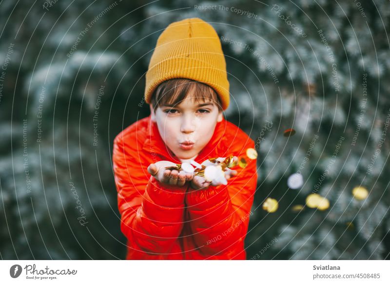 Ein Junge in einer orangefarbenen Jacke und Hut hält glänzendes Konfetti in seinen Händen und pustet darauf, der Junge freut sich über die Feiertage. Urlaubsatmosphäre, Weihnachten, Neujahr