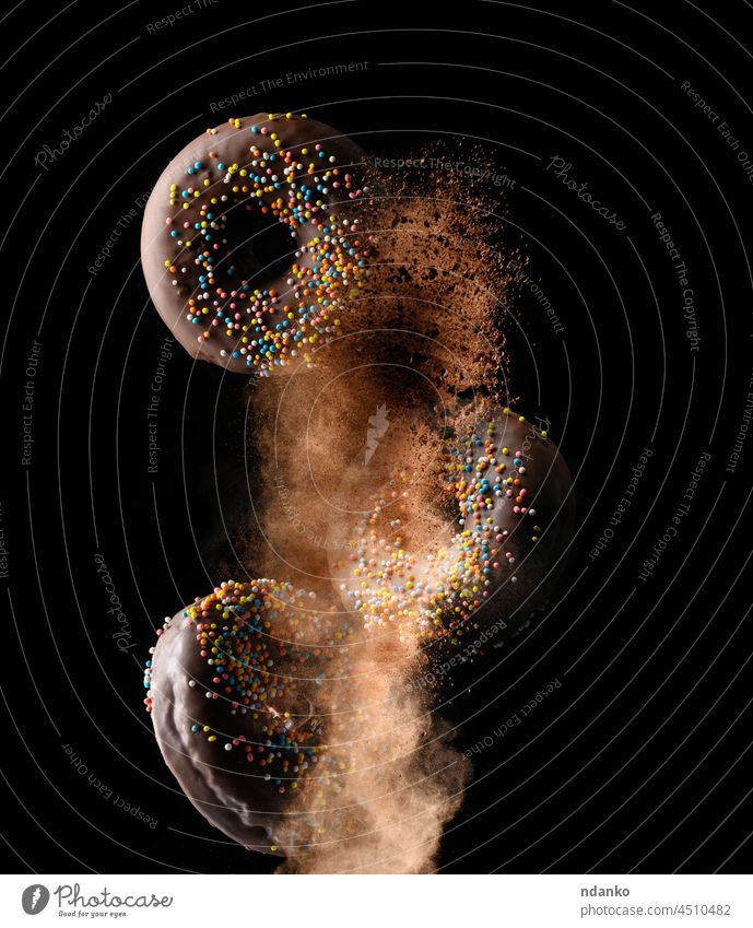 Schokolade runde Donuts mit bunten Zuckerstreuseln schweben in einer Wolke aus braunem Kakao auf einem schwarzen Hintergrund Krapfen Doughnut Lebensmittel süß