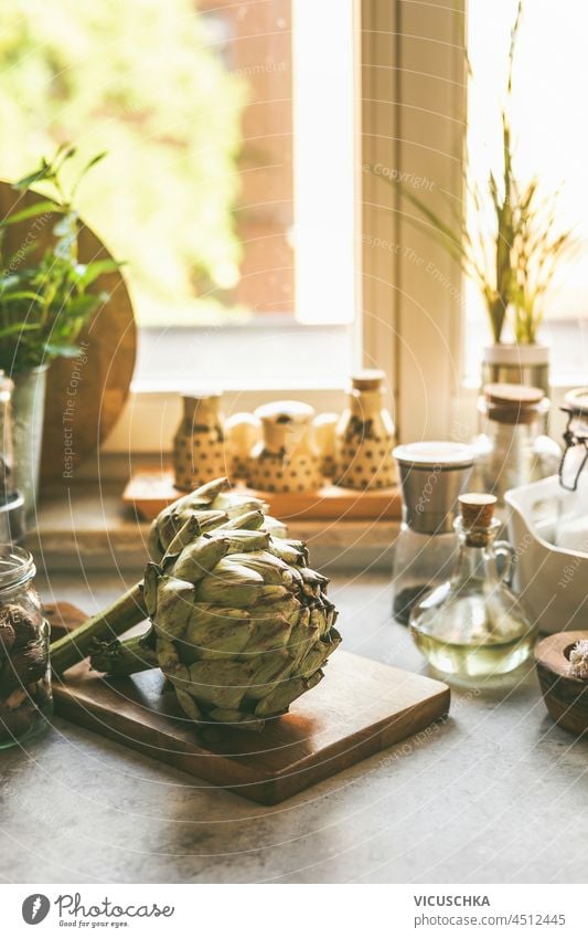 Artischocke auf Küchentisch mit Zutaten am Fenster Hintergrund. Kochen zu Hause mit gesundem und frischem Gemüse. Vorderansicht. Tisch Essen zubereiten