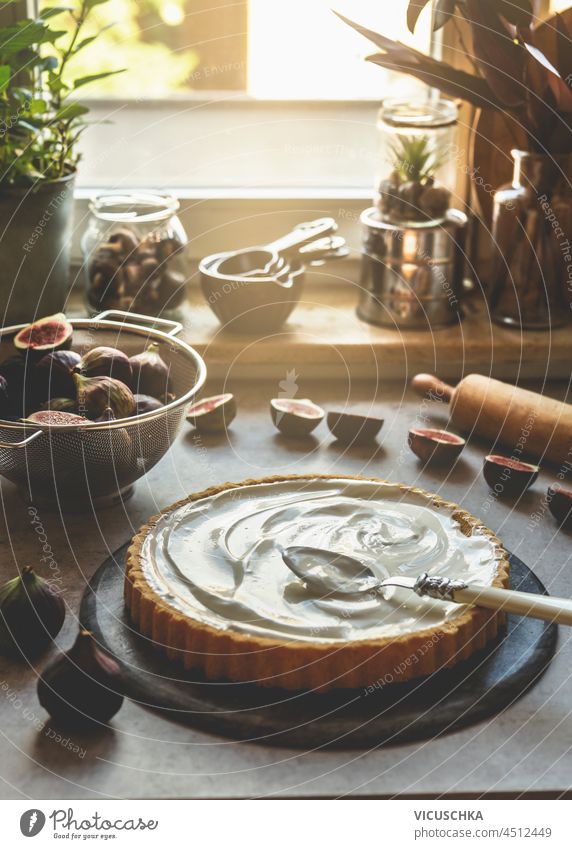 Kuchen mit Creme und Löffel auf dem Küchentisch mit Feigen im Sieb, Nudelholz und Backzubehör im Fensterhintergrund. Backen Vorbereitung zu Hause mit frischen Zutaten. Vorderansicht.