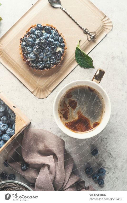 Kaffee in weißer Tasse und Heidelbeerkuchen auf weißem Küchentisch. Kaffeepause zu Hause mit süßem Beeren-Dessert und warmem Getränk. Ansicht von oben.