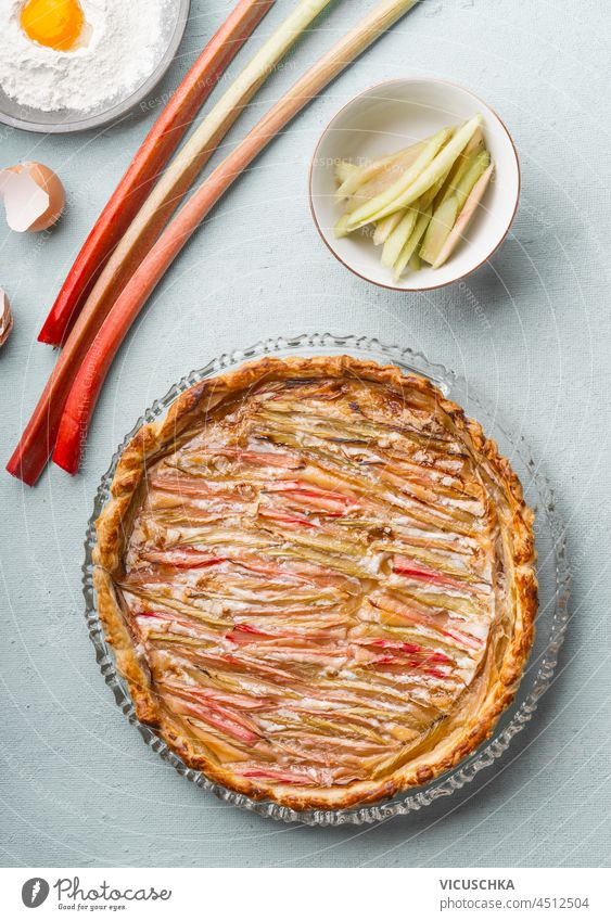 Hausgemachter Rhabarberkuchen auf grauem Küchentisch mit Rhabarberstrauß, Mehl mit Eigelb, Eierschale und Schüssel. Saisonales Backen zu Hause. Ansicht von oben.