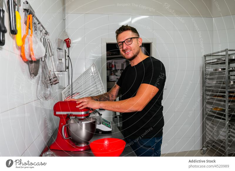 Glücklicher junger Mann in einer Bäckereiküche neben einem Mixer Küche positiv Gebäck Lächeln Konditorei Bestandteil backen Dessert Freude männlich lässig