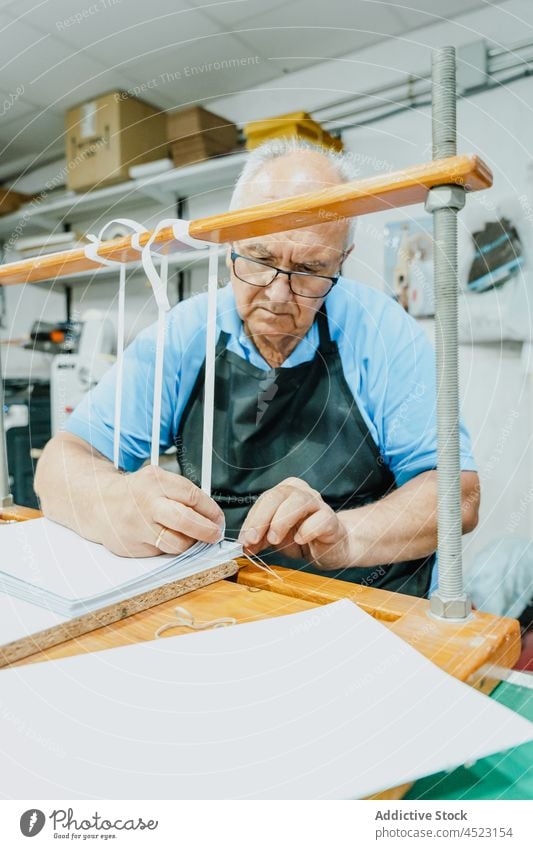 Fokussierter älterer männlicher Handwerker beim Binden von Bändern auf einem Holzbrett im Druckatelier Mann Krawatte Klebeband drucken Druckmaschine Arbeit