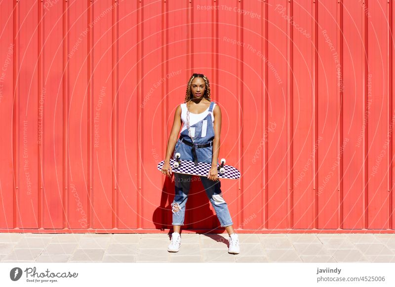 Schwarze Frau gekleidet casual, wtih ein Skateboard auf roten städtischen Wand Hintergrund. schwarz Zopf afrobraid Rastalocken Afrikanisch Frisur Afro-Look