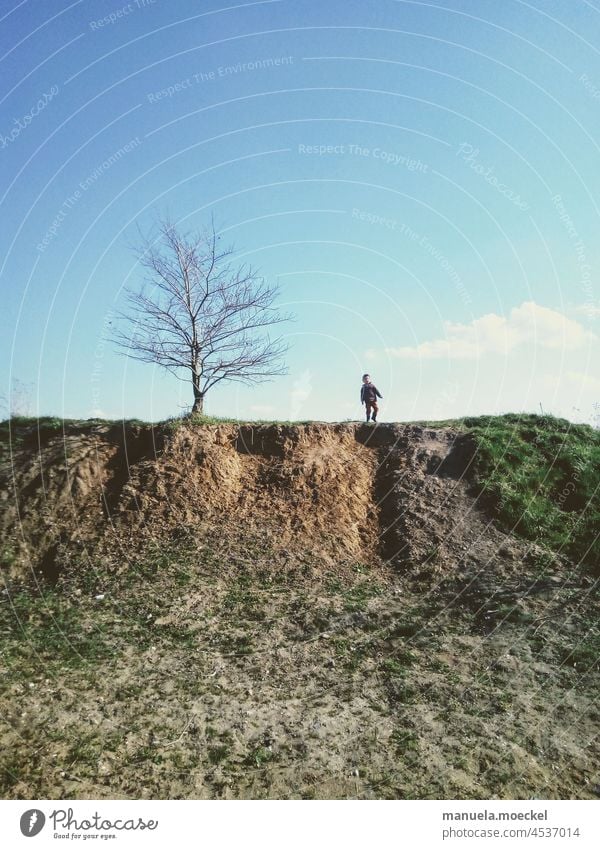 Kleiner Hügel mit Baum und Junge, der ihn erklommen hat. Kind Himmel Wolke braun Erde blau grün Gras