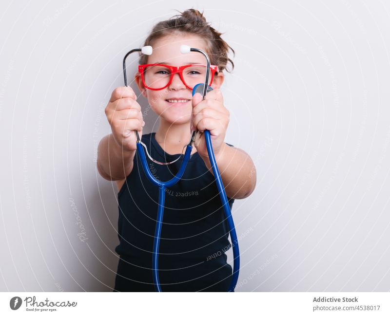 Fröhliches Mädchen im Arzt-Outfit Stethoskop Verkleidung spielen medizinisch spielerisch Kindheit Sanitäter Instrument heiter Glück positiv Atelier Brille