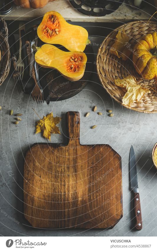 Lebensmittel Hintergrund mit leeren hölzernen Schneidebrett auf Küchentisch mit halbierten Butternusskürbis, Besteck und Küchenutensilien. Herbstliche Kochszene mit saisonalen Zutaten. Ansicht von oben.