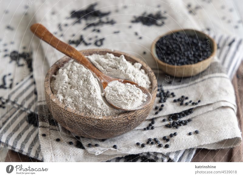 Schüssel mit einem Löffel schwarzem Linsenmehl und Bohnen auf weißem Holztisch Nahaufnahme Mehl Beluga alternativ Hülsenfrüchtler roh getrocknet