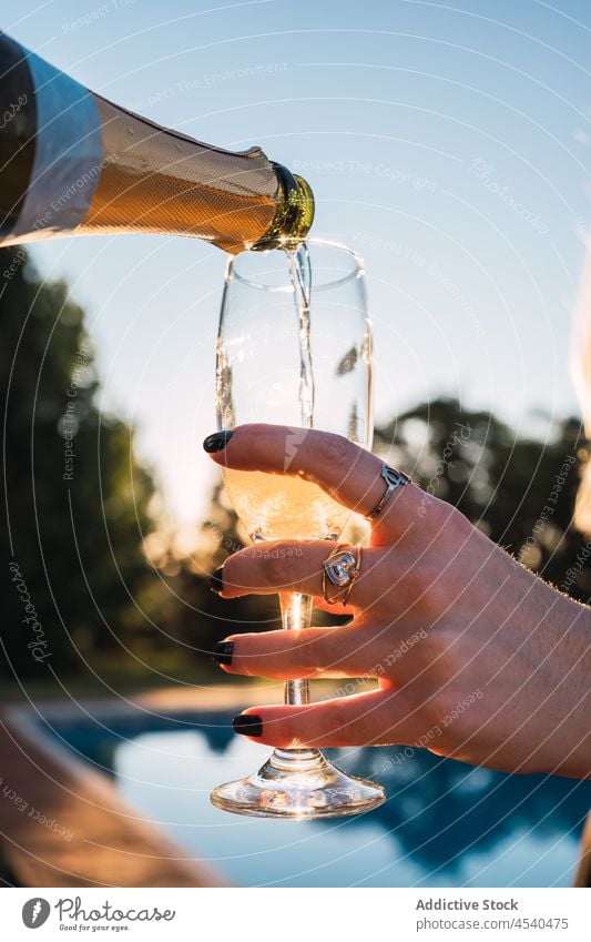 Crop Person mit Champagner in Glas gegossen in der Nähe von Pool eingießen trinken Party Erfrischung Flasche Getränk Schnaps Alkohol Feiertag Beckenrand Resort