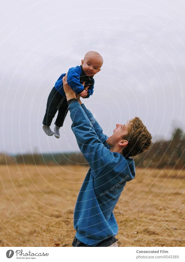 Mann, der sein Baby vor Freude in die Luft hebt, auf dem Lande Säugling neugeboren Junge Babyjunge reisen Abenteuer Lächeln Lachen Glück Liebe Glaube lachen