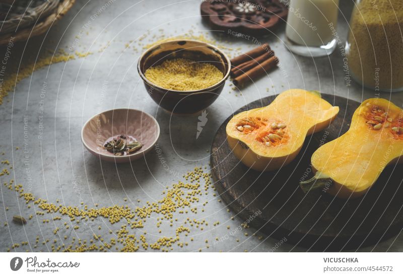 Halbierter Butternusskürbis auf hellgrauem Küchentisch mit Hirse in Schüssel und Küchenutensilien. Zubereitung von gesundem veganem Hirsebrei zu Hause mit saisonalen Herbstzutaten. Ansicht von oben.